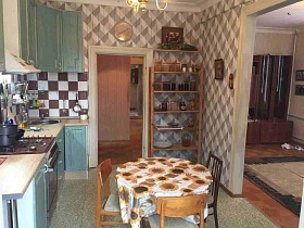 коричневая мебельная горка со стеклянными дверцами в светлой гостиной и прихожая с полосатыми обоями из открытых дверей кухни с зеленой мебельной стенкой и круглым обеденным столом
