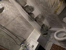 ванная комната с серой плиткой на стенах, серой шторкой над ванной, белым санузлом и коричневым ковриком на полу современной трехкомнатной квартиры