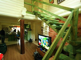 плоский телевизор на тумбе у кирпичной стены под зеленой лестницей напротив черного мягкого дивана в гостиной темной дачи -сруб директора