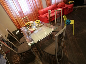 ваза с фруктами, сахарница и стаканы на обеденном стеклянном столе, коричневые стулья с высокой металлической спинкой, желтый детский стульчик на коричневом полу просторной гостиной, совмещенной с кухней квартиры студии молодежной семьи