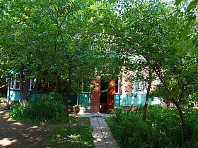 ровная дорожка из плитки между клумбами с густой зеленью под свисающими ветвями деревьев к открытым входным дверям художественной дачи- музей