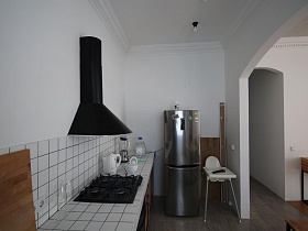 белый детский стульчик для кормления, серебристый холодильник и черная вытяжка над черной газовой плитой в мебельной стенке в зоне кухни современной скандинавской квартиры