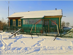 детские ировые снаряды под снегом на участке перед деревянным одноэтажным домом, выкрашенным в зеленый и желтый цвет с верандой посередине
