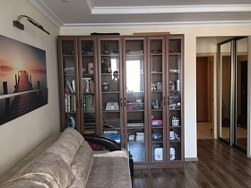 коричневый книжный шкаф со стеклянными дверцами, бежевый мягкий диван у светлой стены с картиной и телевизором гостиной трехкомнатной квартиры