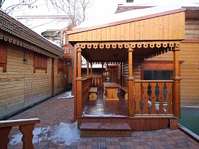 деревянные скамейки у деревянного прямоугольного стола на открытой веранде с резными перилами и наличниками под крышей дома из сруба