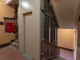 светлая лифтовая кабина за металлической сеткой на лестничной площадке с бежевыми перилами в подъезде, требующего ремонта сталинского дома