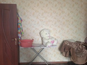 одежда на крючках за открытой коричневой дверью, мягкая игрушка и розовый таз на гладильной доске у светлой стены спальной комнаты квартиры СССР