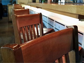 коричневые спинки деревянных барных стульев у барной стойки кафе в темных тонах с большими окнами