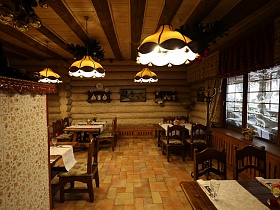 посуда на коричневых полках, картины на бревенчатых стенах, желтые абажуры подвесных люстр на двухцветном деревянном потолке над столиками в уютном зале ресторана в купеческом стиле
