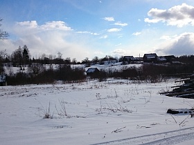 общий вид заброшенной деревни с деревянными домами в снегу на пригорке и грудой бревен у дороги