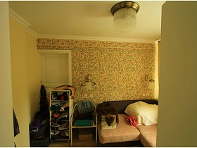 вещи на открытых полках невысокой этажерки, стул, бежево коричневый угловой диван у стены с цветочными обоями и круглой люстрой на потолке