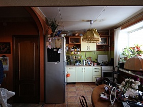 комнатный цветок на верху серебристого холодильника, кухонная утварь на столешнице белой мебельной стенки с навесными коричневыми шкафами под стеклом и открытыми полками у стены кухни с большим окном