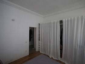 белые шторы на встроенном шкафу с открытыми полками, шкафчиками и одеждой на тремпелях в светлой спальне скандинавской квартиры