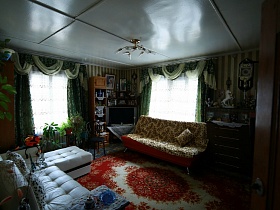 белый угловой диван и раскладной диван на красном цветном ковре гостиной с многочисленными статуэтками, сувенирами в шкафу, на комоде деревянной избы в деревне