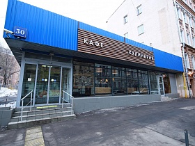 фасад здания уличного кафе на высоком фундаменте с лестницей и металлическими перилами у главного входа с раздвижными дверьми,большими окнами, видеокамерой на голубой крыше