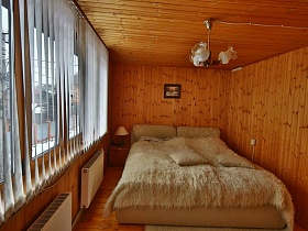 люстра над большой кроватью с меховым покрывалом и прикроватная тумбочка с торшером у большого окна с белыми жалюзи  в спальне загородной дачи