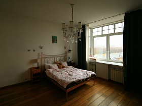 настольные лампы на деревянных прикроватных тумбочках у большой кровати с цветным постельным на коричневом полу спальной комнаты с белой гардиной и черными шторами на окне