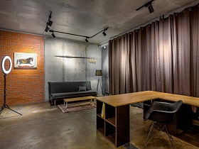 картина на стене из красного кирпича слева от серого дивана, торшер на ножках - справа в стильной студии с коричневыми шторами на окнах