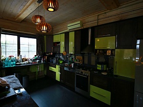 коричневая с салатовым мебельная кухня со встроенной газовой плитой, мультиваркой, электрочайником и прочими кухонными предметами на столешнице у стены гостиной современного деревянного загородного дома