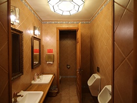 бра и прямоугольные зеркала в рамке на стене с бежевой плиткой над белыми раковинами в деревянной столешнице туалетной комнаты русского ресторана