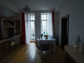 белая гардина и красные шторы на высоком окне и балконной двери белой кухни с обеденным столом посередине и коричневой мебельной стенкой
