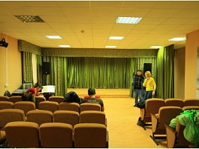 актовый зал с рядами бежевых мягких кресел, зеленым занавесом на сцене школы в деревне