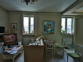 картины на полу за журнальным столиком у мягкого светлого дивана, картина на мольберте в углу гостиной и картина на стене между окнами со ставнями в современной дизайнерской квартире с видом на Москву реку