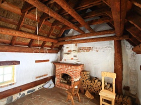 поленница у камина из кирпича, деревянные стулья внутри дома мазанки с белыми стенами под высокой крышей из дерева