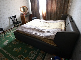 черные стулья со спинкой, черная мягкая кровать с подушками и полосатым покрывалом, столик у окна с коричневыми шторами светлой спальной комнаты простой современной квартиры