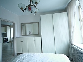 белый двухдверный шкаф для одежды у большого окна, прямоугольное зеркало над белой высокой тумбой у открытой двери спальной комнаты двухкомнатной квартиры