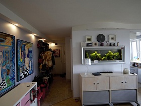 настенный аквариум, часы и картинки в белой рамке на стене над низким двухцветным шкафом в светлой гостиной современной трехкомнатной квартиры в Икеа стиле