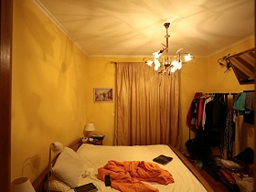 телевизор и бра на желтой стене, над напольными вешалками с одеждой, настольные лампы на тумбочках у большой деревянной кровати в спальной комнате дизайнерской квартиры