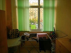 столик,сушилка для белья, комод, полочка у окна застекленной лоджии с зелеными  жалюзи простой квартиры молодоженнов