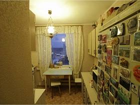 многочисленные магнитики на дверце холодильника, белый плафон подвесной люстры на потолке кухни с мебельной стенкой, обеденным столом у окна с белыми шторами квартиры СССР 80-89 гг стиля