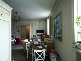 картины, белый плоский телевизор на светлой полосатой стене с открытой дверью в спальную комнату из зоны кухни девчачьей дизайнерской квартиры