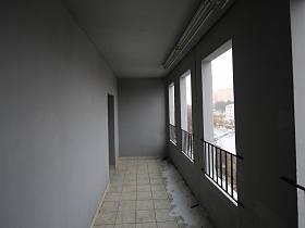 просторная белая лоджия с квадратной плиткой на полу и металлическими перилами на окнах современной многоэтажки в новостройках