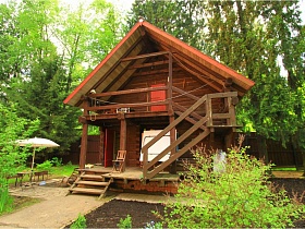 небольшой деревянный двухэтажный домик с деревянными лестницами и открытым балконом под треугольной крышей на участке современного дома для съемок кино