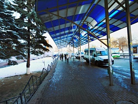 автобусная остановка с тротуарной плиткой на площадке, невысоким металлическим забором под яркой бело синей полосатой крышей у высоких зеленых елей в зимнее  время