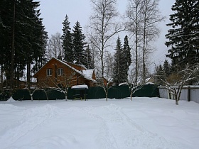 большой участок кирпичного дома в снегу в окружении высоких зеленых елей и соседних домов