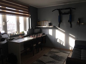 спортивный тренажер на серой стене юношеской комнаты с серым мягким диваном, серым ковром на полу, письменными столами у большого окна с жалюзи трехкомнатной квартиры