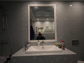 строго и лаконично в ванной комнате- прямоугольное зеркало в белой рамке на стене с серой плиткой над белой раковиной в загородном домике