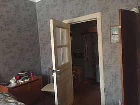 стул со спинкой, прикроватная тумбочка за открытой дверью спальной комнаты со шкафом у стен с серыми обоями с цветочным рисунком