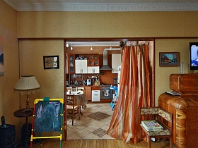 настольная лампа на круглом деревянном столике, детская доска для рисования, цветное полукресло у дверного проема с ораньжевой шторой в гостиной трехкомнатной квартиры эпохи СССР