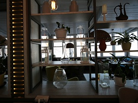 стеклянные вазы, восточные кувшины, комнатные цветы на деревянных полках открытого стеллажа по центру светлого зала стильного ресторана