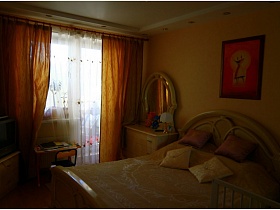 зеркало с комодом и кровать со спинкой цвета слоновой кости у стены с яркой картиной в спальне приличной трешки панельного дома