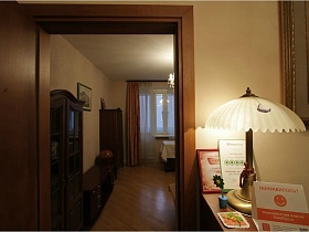 бронзовый торшер с белым абажуром, вязанная салфетка, исскуственный цветок, грамота в рамке на ресепшн у входной двери в спальную комнату современной трехкомнатной съемной квартиры