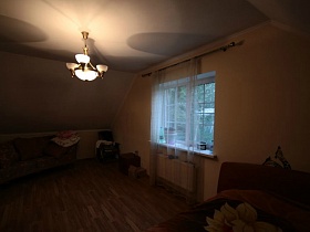 угловой диван с подушками, кресло и прикроватная тумбочка у окна с белой гардиной в спальне на мансарде простого семейного дома в густом лесу