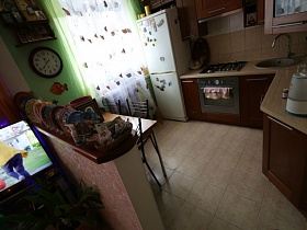 полочка с иконами, круглые часы на стене над обеденным столиком в зоне кухни с бежевой плиткой на полу в комнате в областном квартале