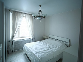 подвесная люстра с белыми стеклянными плафонами на белом потолке светлой спальной комнаты с белыми шторами на большом окне стильной квартиры