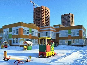 разноцветный двухэтажный детский сад с разнообразными яркими деревянными домиками, паравозиками, качелями на заснеженном участке внутри жилого квартала новостройки с высотками